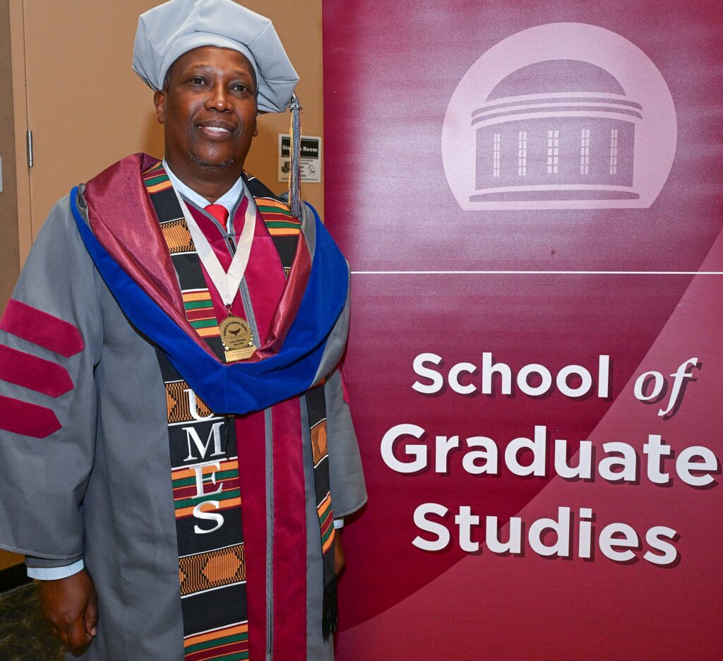 UMES doctoral graduate John Ithiru is pictured by the School of Graduate Studies banner at UMES' spring 2026 commencement