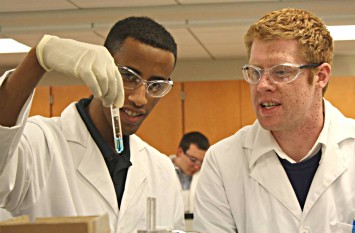 Students wearing goggles and lab coats discuss the contents of a test tube. 